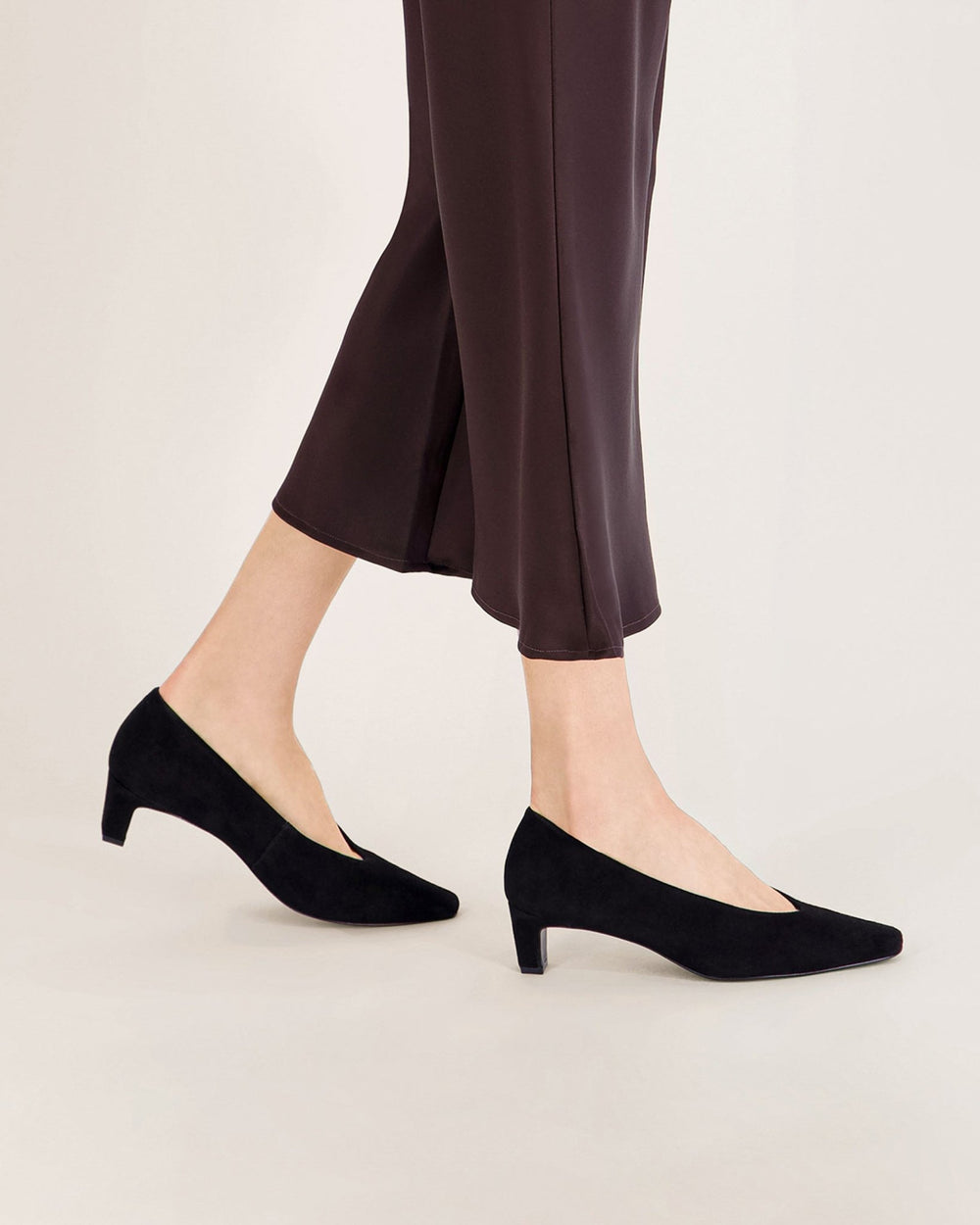 Black suede kitten heel pumps and dark brown knee-high boots on a wood floor beside a folded wool coat