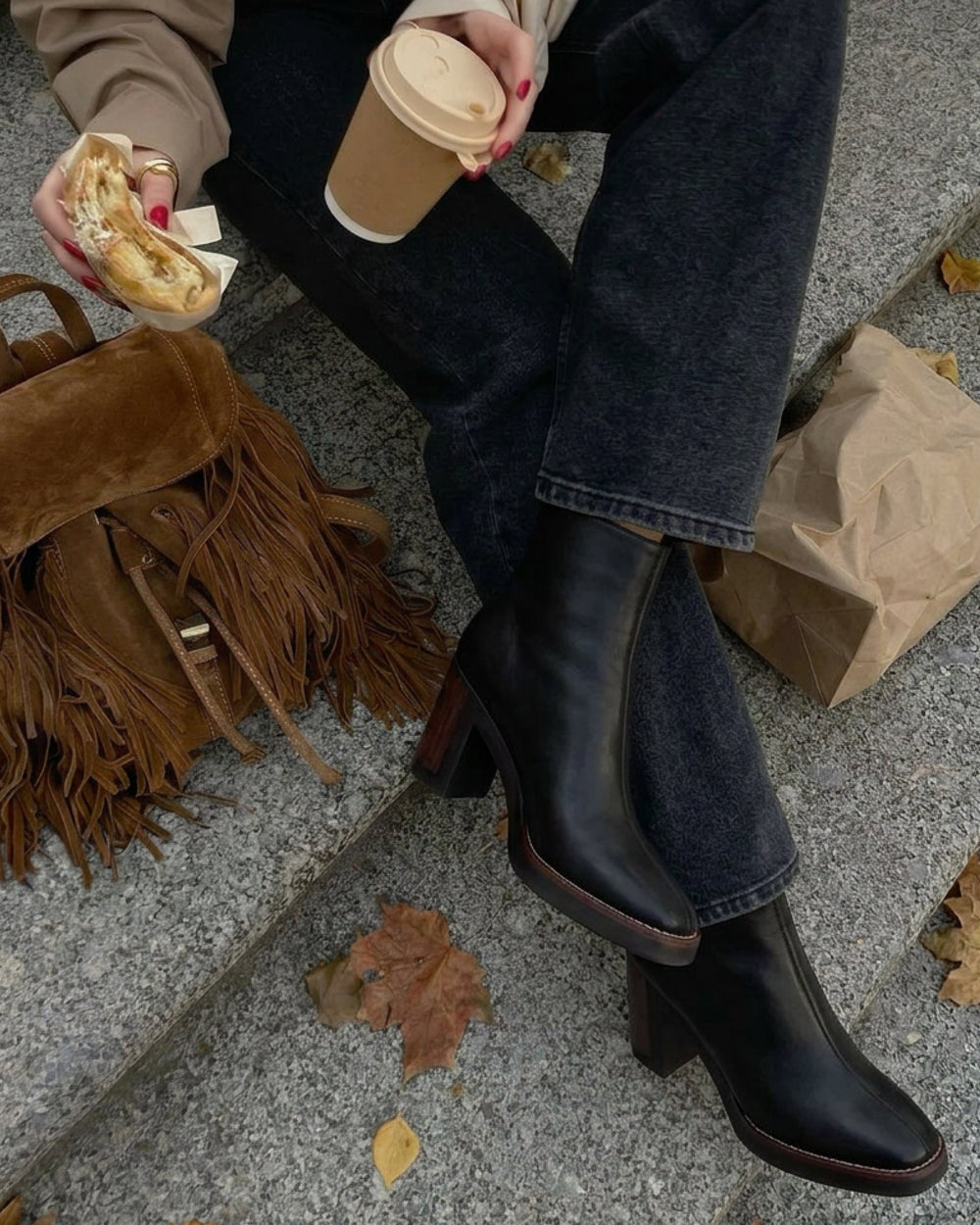 Black Nara Platform Booties with straight dark jeans, a trench over a cream shirt, coffee in hand, fringed suede backpack, on stone steps with fall leaves.